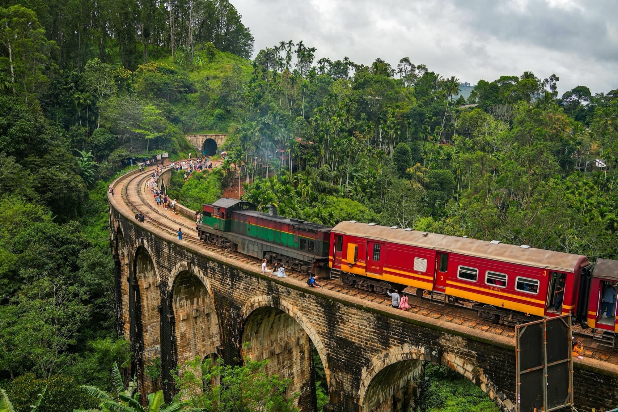 A red train crossing a stone viaduct through hill-country forest