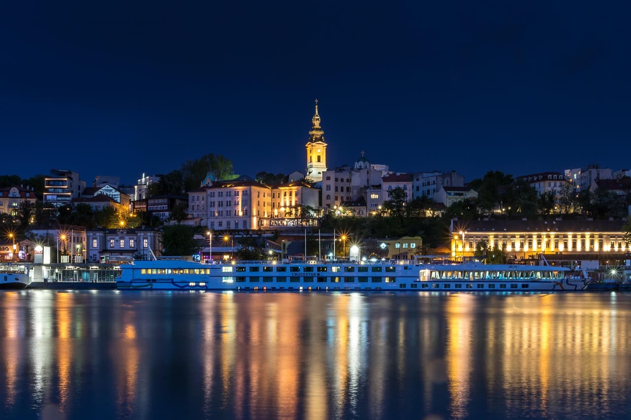 A night landscape of Belgrade's illuminated skyline and a cathedral reflected in the calm waters of the Sava River with a cruise ship docked.
