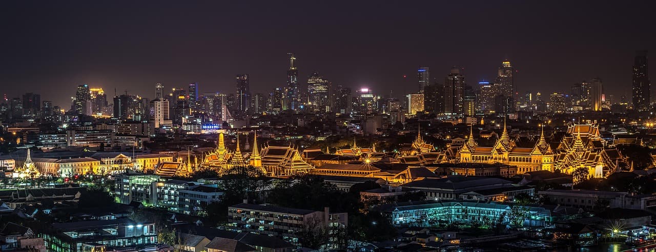A wide panoramic view of Bangkok at night, showing illuminated traditional temples in the foreground and a modern city skyline in the distance.