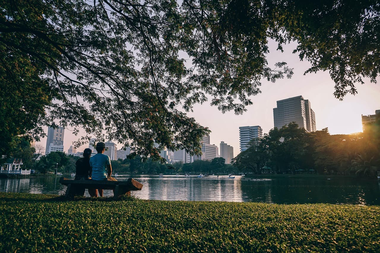 Two people sitting on a bench in a green park, looking across a calm lake toward a city skyline at sunset.