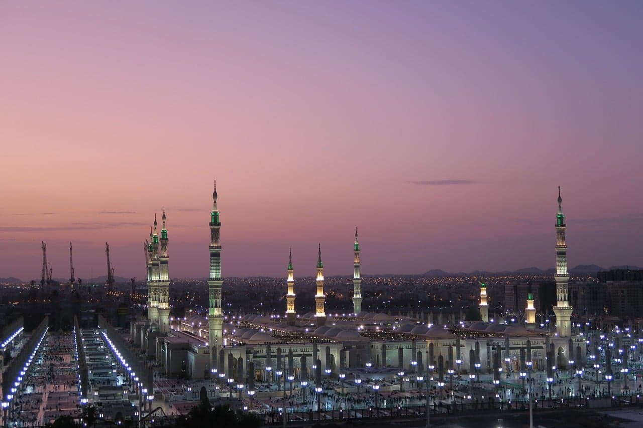 A wide shot of a large illuminated mosque with multiple minarets at dusk in Saudi Arabia.