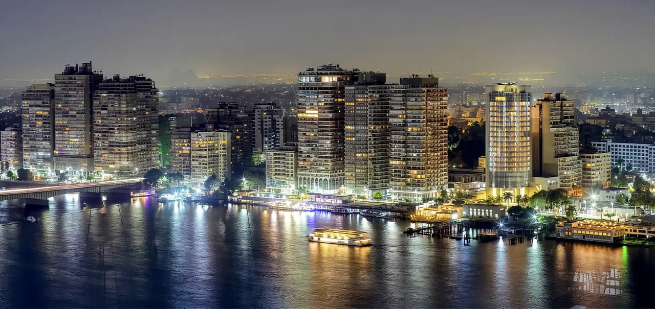 A night view of Cairo's skyline along the Nile River, with illuminated buildings and a boat reflected in the dark water.