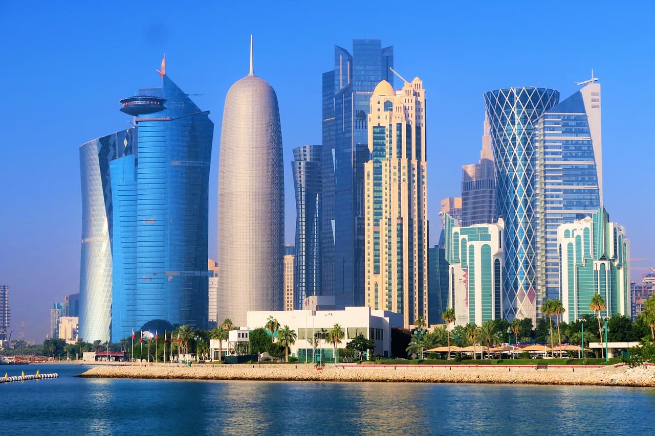 A vibrant landscape photo of the Doha skyline in Qatar featuring modern skyscrapers against a clear blue sky and water in the foreground.
